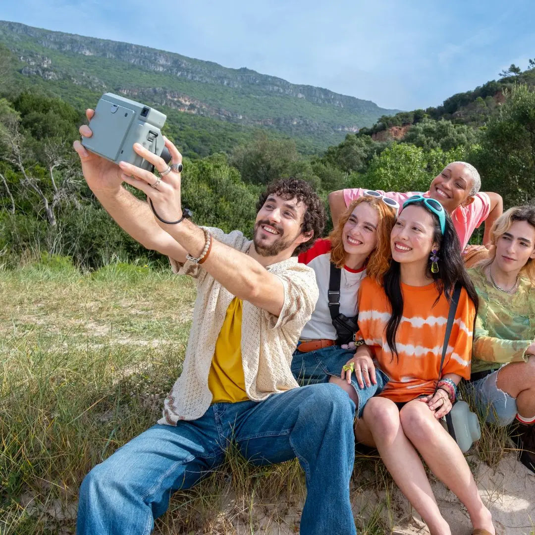 Group of friends taking a selfie using Instax Wide 400 in a scenic outdoor setting with mountains and greenery.