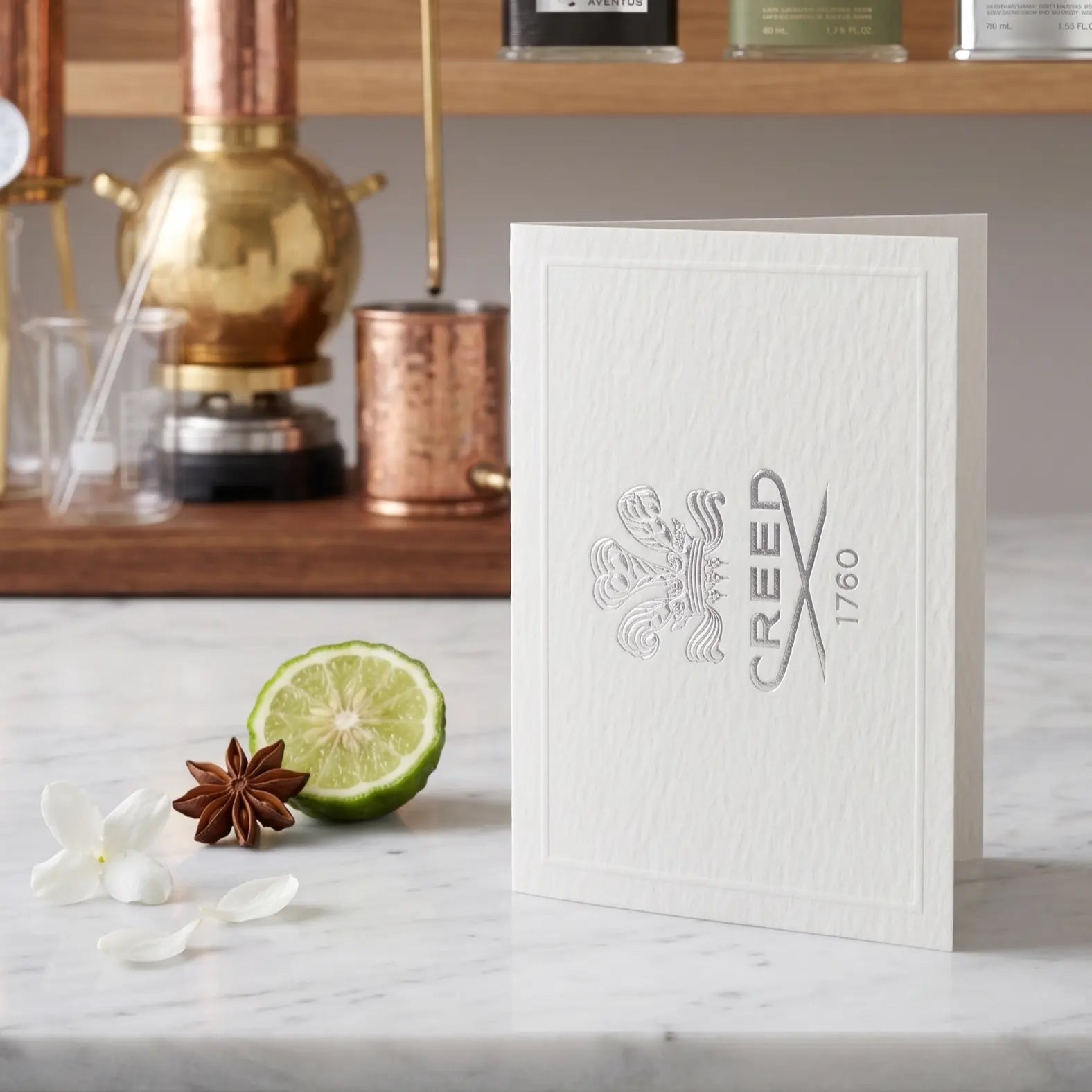 Creed vial with lime, star anise, and flowers on a marble surface with a distillation setup in the background.